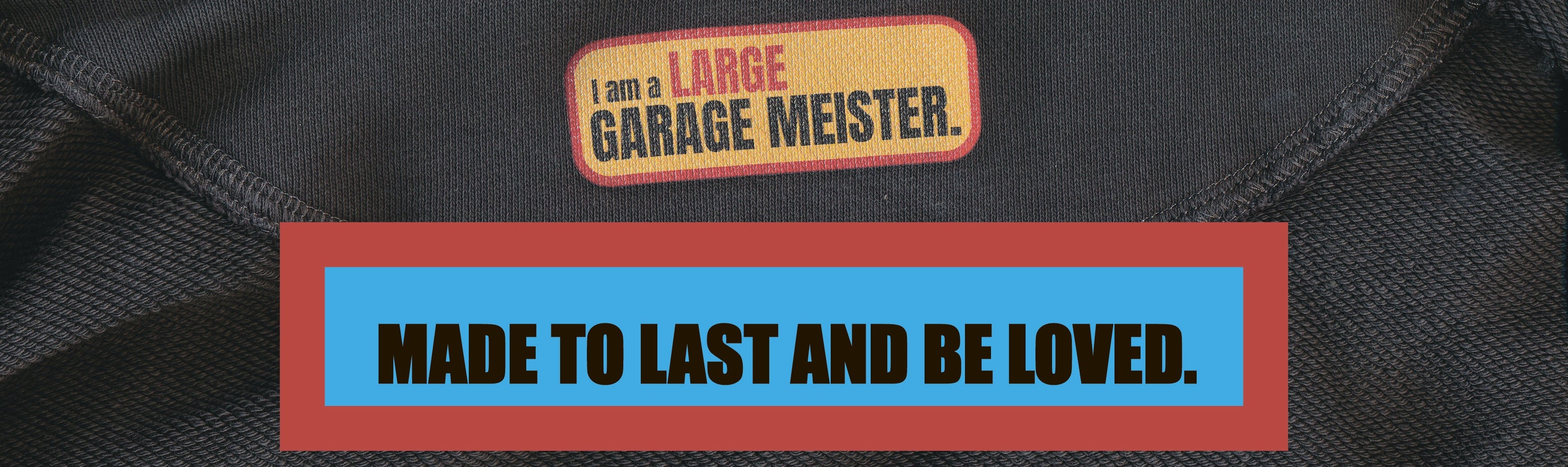 A close-up of a black garment with a small rectangular patch reading 'I am a LARGE GARAGE MEISTER.' above a large red-bordered blue text panel that says 'MADE TO LAST AND BE LOVED.